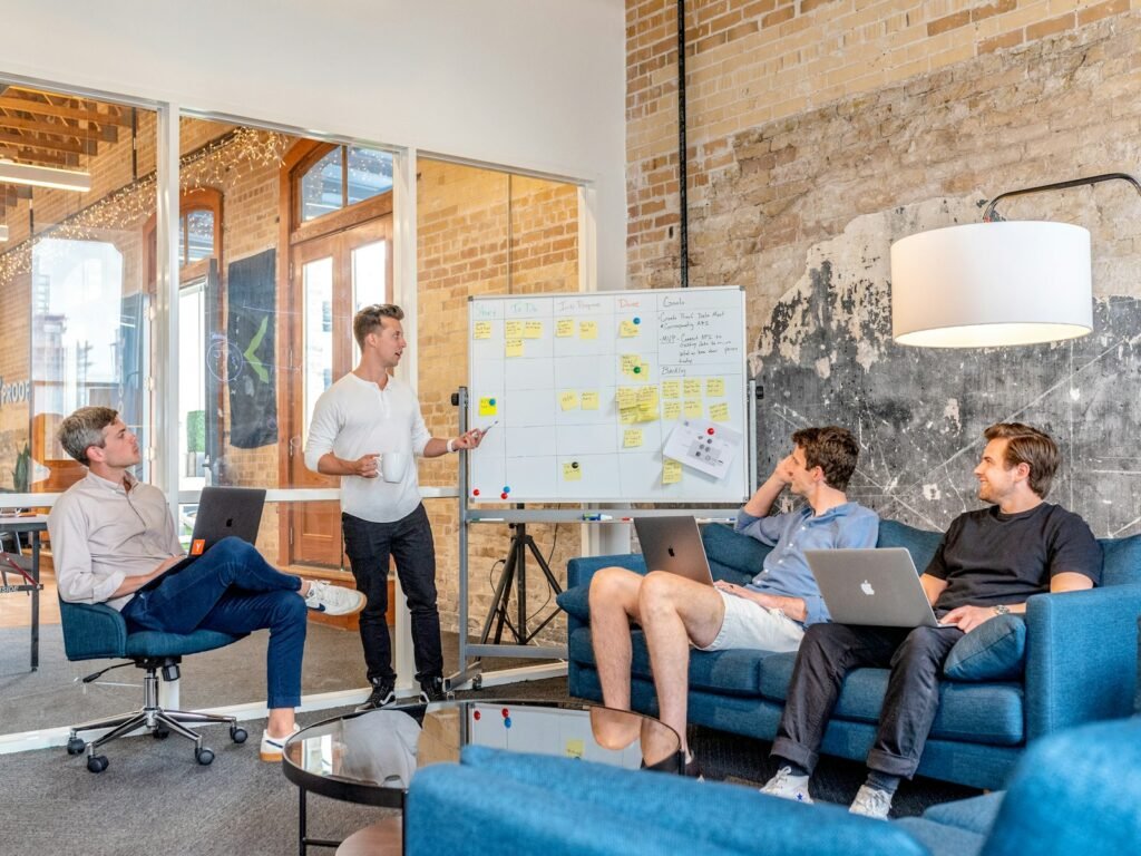 Three men sitting while using laptops and watching man beside whiteboa